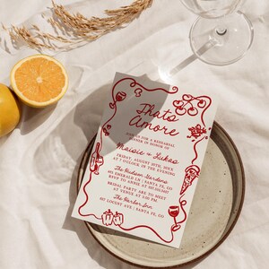 May include: A red and white invitation card with the text "Thato Amore" and details for a rehearsal dinner in honor of Maisie and Lukas. The invitation includes the date, time, location, and RSVP information. The card is decorated with red hearts, cherries, wine glasses, and a pizza slice.