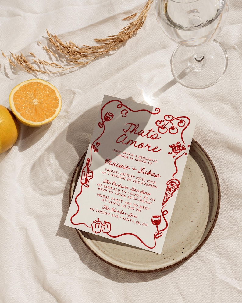 May include: A red and white invitation card with the text "Thato Amore" and details for a rehearsal dinner in honor of Maisie and Lukas. The invitation includes the date, time, location, and RSVP information. The card is decorated with red hearts, cherries, wine glasses, and a pizza slice.