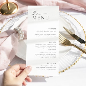 May include: A menu card with the text "The Menu" in elegant script. The card lists starters, entrees, and desserts. The menu is held over a white plate with gold cutlery and a pink napkin.