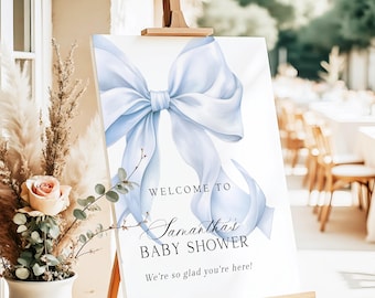 May include: A white sign on a wooden easel with a large, light blue bow illustration. Text reads "WELCOME TO Samantha's BABY SHOWER. We're so glad you're here!" Decorative floral arrangement in a white vase is in the foreground.