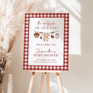 Puede incluir: Un letrero de bienvenida para baby shower con un borde de cuadros rojos y blancos. El letrero presenta el texto "The Apple of Our Eye!" y "Welcome to Samantha's Baby Shower." Elementos decorativos: manzanas y ropa de beb&eacute;.