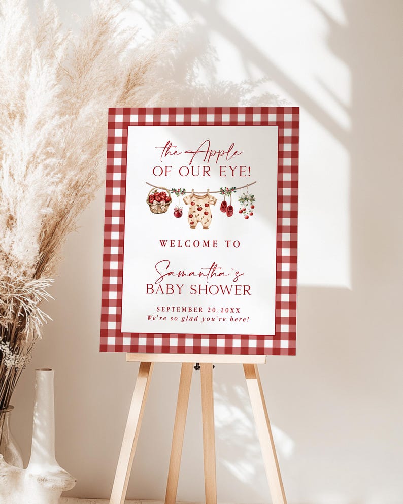 Puede incluir: Un letrero de bienvenida para baby shower con un borde de cuadros rojos y blancos. El letrero presenta el texto "The Apple of Our Eye!" y "Welcome to Samantha's Baby Shower." Elementos decorativos: manzanas y ropa de beb&eacute;.