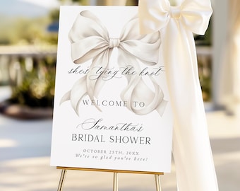 May include: A white rectangular bridal shower welcome sign on a gold easel, featuring a large illustration of a cream-coloured ribbon bow. The text reads: she's tying the knot, WELCOME TO, Samantha's BRIDAL SHOWER, OCTOBER 25TH, 20XX, We're so glad you're here!