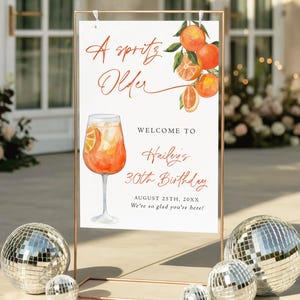 May include: A sign with the text "A Spritz Older" and "Welcome to Hailey's 30th Birthday". The sign features a watercolor illustration of an orange cocktail and oranges. The sign is displayed in a copper-colored frame, with disco balls in the foreground.