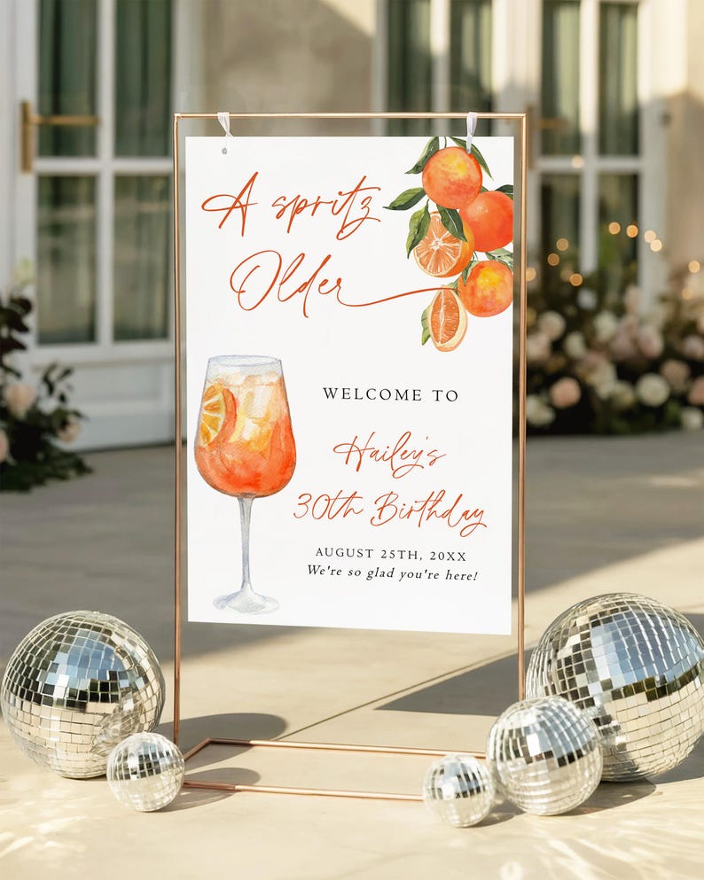 May include: A sign with the text "A Spritz Older" and "Welcome to Hailey's 30th Birthday". The sign features a watercolor illustration of an orange cocktail and oranges. The sign is displayed in a copper-colored frame, with disco balls in the foreground.