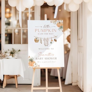 May include: A white rectangular welcome sign for a baby shower on a wooden easel. The sign features watercolor illustrations of pumpkins, a clothesline with a baby onesie, and text reading: A little PUMPKIN IS ON THE WAY! WELCOME TO Samantha's BABY SHOWER We're so glad you're here!