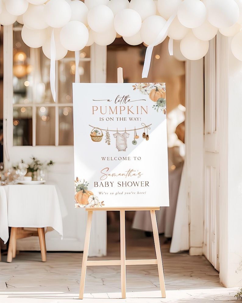 May include: A white rectangular welcome sign for a baby shower on a wooden easel. The sign features watercolor illustrations of pumpkins, a clothesline with a baby onesie, and text reading: A little PUMPKIN IS ON THE WAY! WELCOME TO Samantha's BABY SHOWER We're so glad you're here!