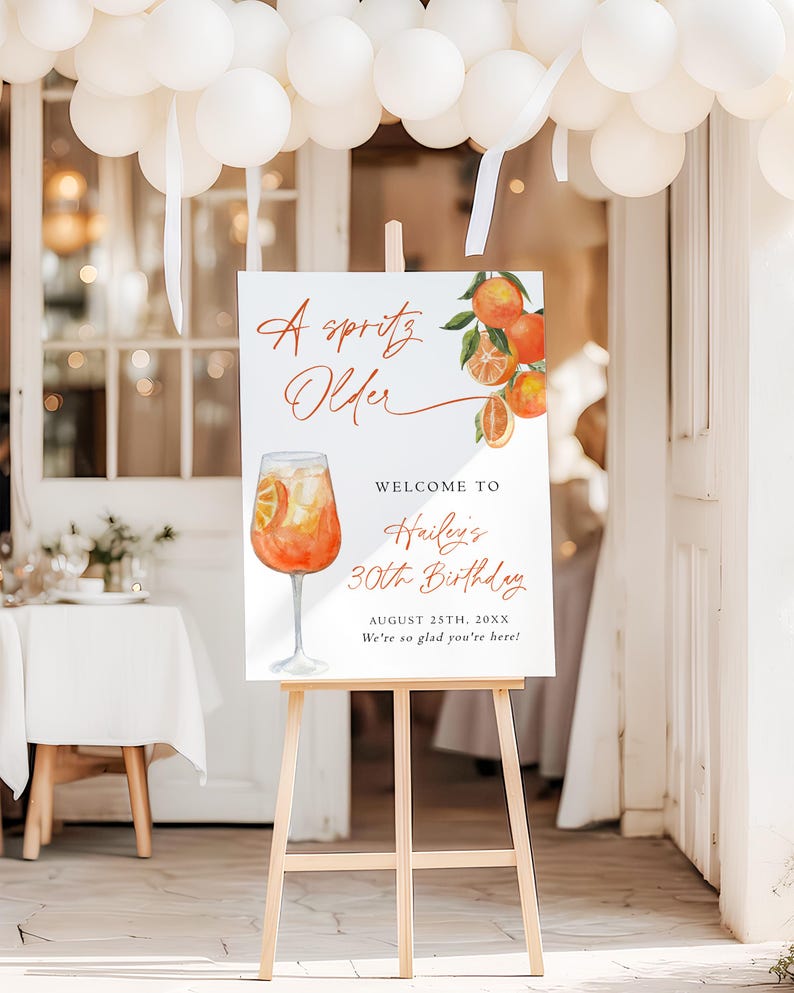 May include: A white sign on a wooden easel with the text "A Spritz Older" and "Welcome to Hailey's 30th Birthday". The sign features an illustration of an orange cocktail and orange slices.
