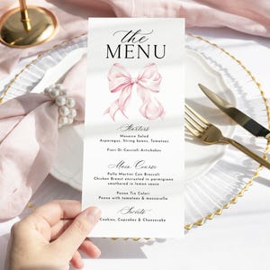 May include: A menu card with the text "The MENU" and a watercolor bow illustration. The menu lists starters, main course, and sweets. The card is held over a white plate with gold cutlery and a pink napkin.
