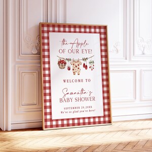 Puede incluir: Un letrero enmarcado para baby shower con un borde a cuadros rojo y blanco. El letrero dice "the Apple of our Eye!" y "Welcome to Samantha's Baby Shower" con la fecha. El dise&ntilde;o incluye ilustraciones de manzanas y ropa de beb&eacute;.