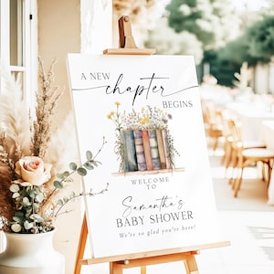 May include: A white sign with a watercolor illustration of a stack of books with flowers. The text reads "A new chapter begins. Welcome to Samantha's Baby Shower. We're so glad you're here!"