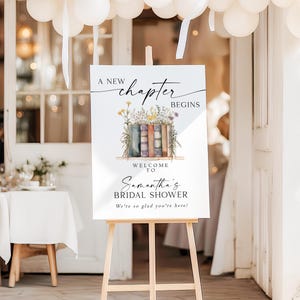 May include: A white sign on a wooden easel with the text "A New Chapter Begins" and "Samantha's Bridal Shower." The sign features a watercolor illustration of books and flowers. White balloons are suspended above.