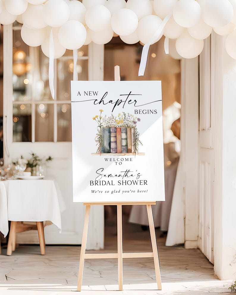 May include: A white sign on a wooden easel with the text "A New Chapter Begins" and "Samantha's Bridal Shower." The sign features a watercolor illustration of books and flowers. White balloons are suspended above.
