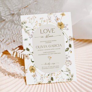 K&ouml;nnte beinhalten: Eine florale Brautduscheinladung mit einem Aquarelldesign aus gelben und gr&uuml;nen Blumen. Der Text lautet "Love in Bloom" und enth&auml;lt den Namen der Braut, Datum, Uhrzeit und Ort. Die Einladung enth&auml;lt auch die RSVP-Informationen.