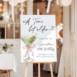 May include: A white sign on a wooden easel with the text "A Tini bit older" and "Welcome to Samantha's 30th Birthday." A martini glass with olives and a pink bow is illustrated below the text. White balloons are in the background.