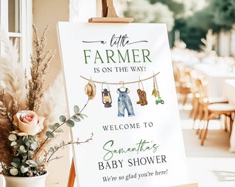 May include: A white sign on an easel with the text "a little FARMER is on the way!" in green and black. The sign welcomes guests to Samantha's baby shower. Illustrations of farm-themed items hang from a clothesline.