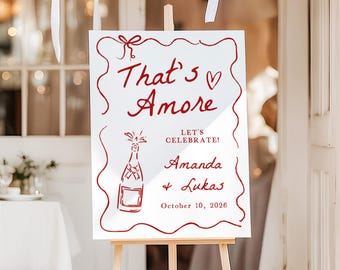 May include: A white sign with red text and a decorative border, displayed on a wooden easel. The sign reads "That's Amore" and includes the names Amanda and Lukas, along with the date October 10, 2026. White balloons are visible in the background.