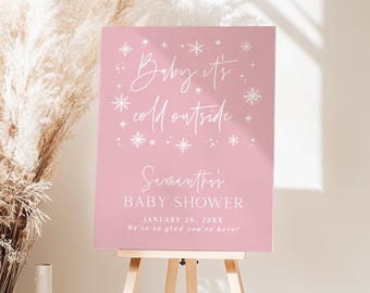 May include: A pink baby shower sign on a wooden easel. The sign has white text that reads "Baby it's cold outside" and "Samantha's BABY SHOWER January 20, 20XX We're so glad you're here!" Decorative snowflakes are also included.