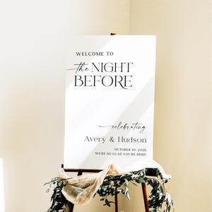 Pu&ograve; includere: Un cartello di benvenuto per matrimoni bianco su un cavalletto di legno. Il cartello recita "WELCOME TO the NIGHT BEFORE celebrating Avery & Hudson OCTOBER 10, 2026 WE'RE SO GLAD YOU'RE HERE!". Il cartello &egrave; decorato con verde e tessuto.