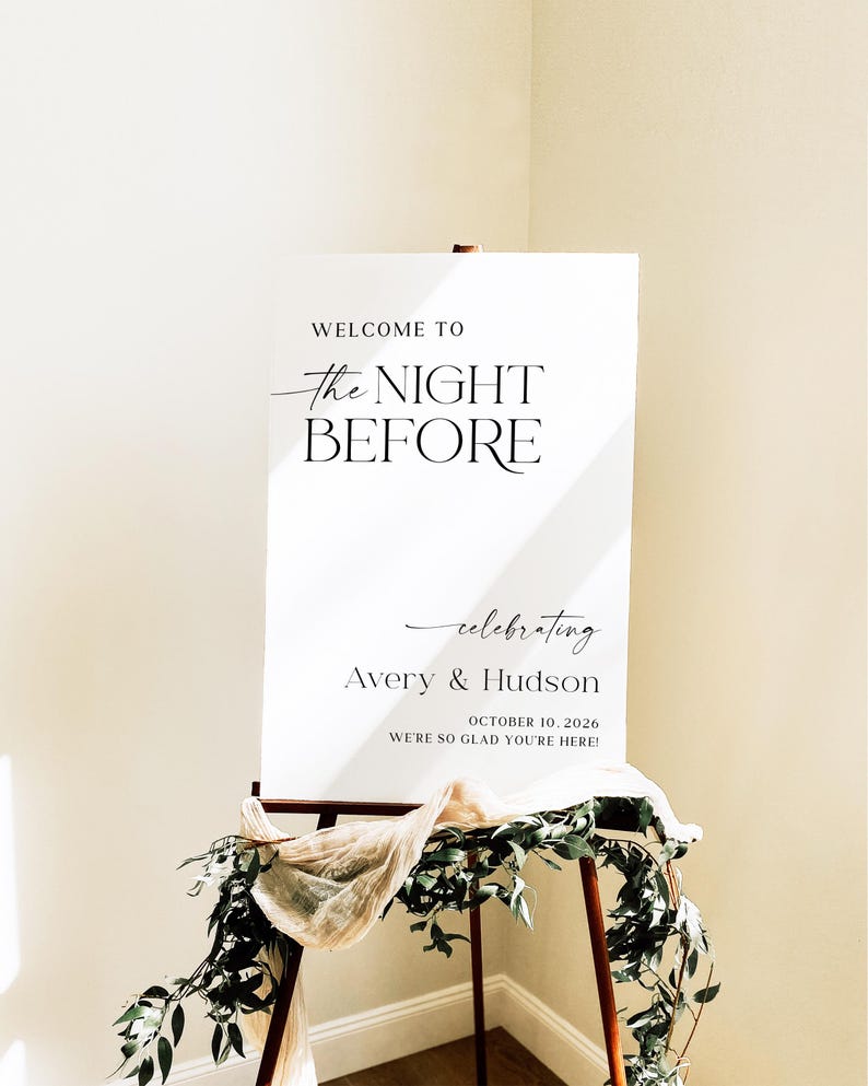 Pu&ograve; includere: Un cartello di benvenuto per matrimoni bianco su un cavalletto di legno. Il cartello recita "WELCOME TO the NIGHT BEFORE celebrating Avery & Hudson OCTOBER 10, 2026 WE'RE SO GLAD YOU'RE HERE!". Il cartello &egrave; decorato con verde e tessuto.