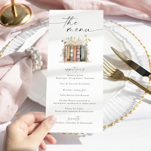 May include: A menu card with the text "the menu" in elegant script. The card features a watercolor illustration of books and flowers. The menu lists appetizers, main course, and sweets. Gold cutlery and a white plate are also visible.