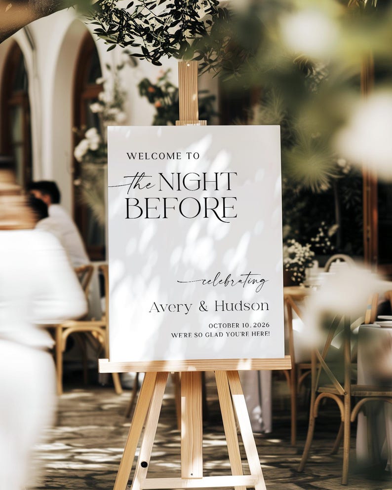 Pu&ograve; includere: Un cartello bianco su un cavalletto di legno con la scritta "WELCOME TO the NIGHT BEFORE celebrating Avery & Hudson OCTOBER 10, 2026 WE'RE SO GLAD YOU'RE HERE!" in nero. Il cartello &egrave; all'aperto.