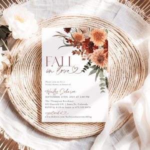 May include: A bridal shower invitation with the words "FALL in love" in a burgundy script. The invitation features floral arrangements in shades of orange, burgundy, and green. The text includes the date, time, and location of the event.