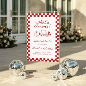 May include: A rectangular event sign with a red and white checkered border, featuring the text That's Amore!, Let's Celebrate!, IN HONOR OF, Maddie & Lukas, AUGUST 16TH, 2025, SANTA FE, COLORADO, and red line art of pizza and wine.