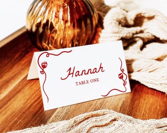 May include: A white folded place card featuring red decorative line art of wine glasses and swirling vines. The text Hannah is printed in a red script font, with the words TABLE ONE printed below in a smaller, simple red serif font.