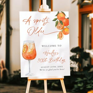 May include: A white sign on a wooden easel with the text "A Spritz Older" and "Welcome to Hailey's 30th Birthday". The sign features watercolor illustrations of oranges and a cocktail. The date is August 25th, 20XX.