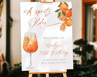 May include: A white sign on a wooden easel with the text "A Spritz Older" and "Welcome to Hailey's 30th Birthday". The sign features watercolor illustrations of oranges and a cocktail. The date is August 25th, 20XX.
