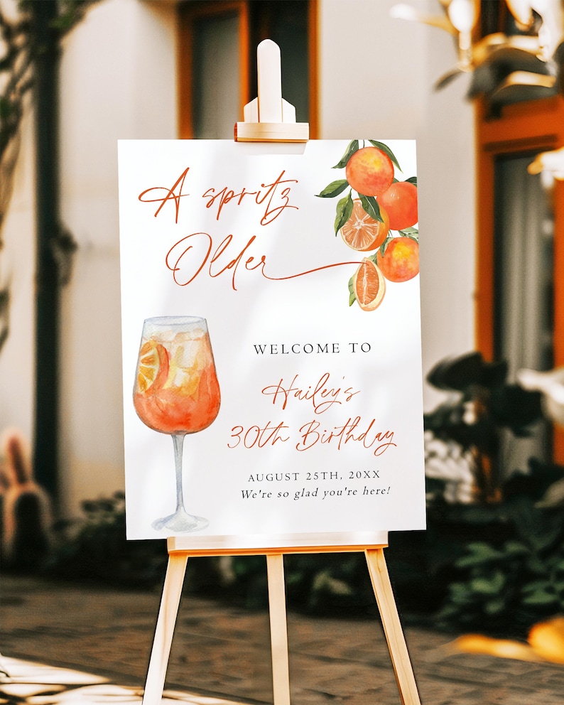 May include: A white sign on a wooden easel with the text "A Spritz Older" and "Welcome to Hailey's 30th Birthday". The sign features watercolor illustrations of oranges and a cocktail. The date is August 25th, 20XX.