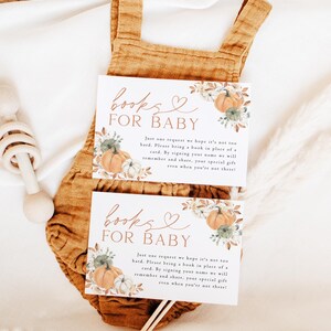 May include: Two baby shower invitation cards with the text "Books for Baby" and a request for a book in place of a card. The cards feature pumpkin and floral illustrations. They rest on a mustard-colored baby romper.
