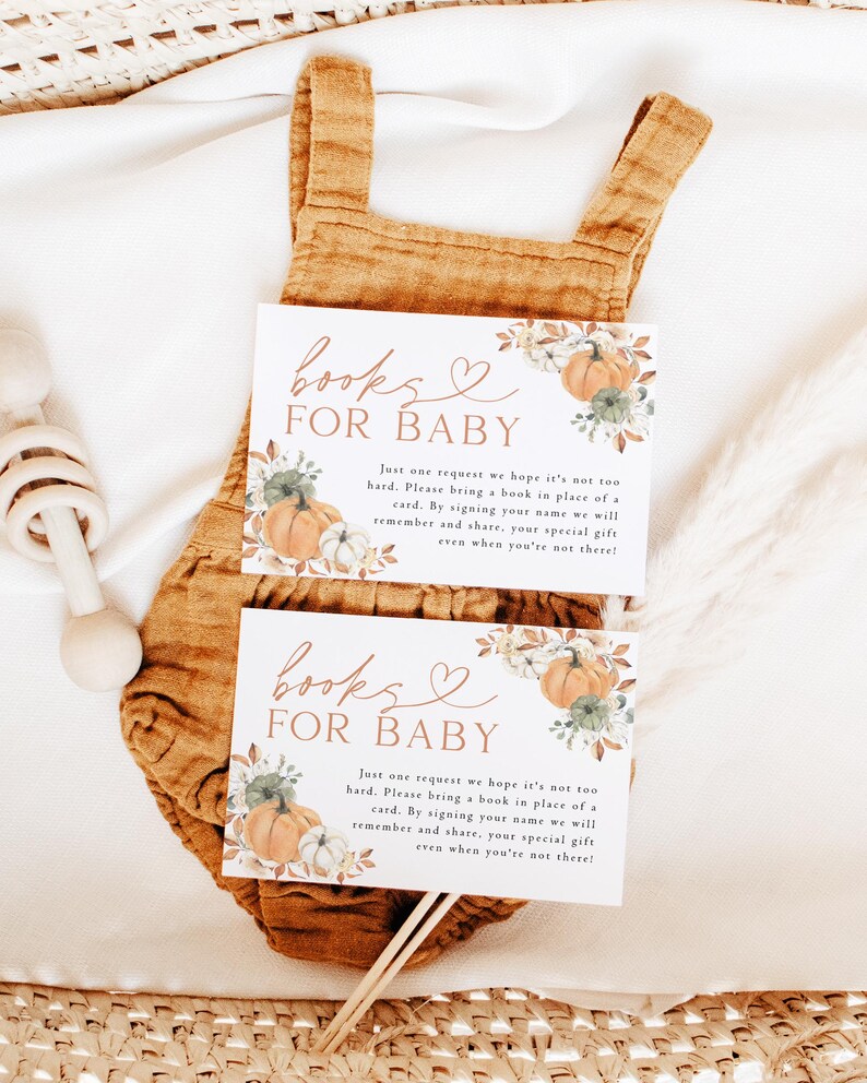 May include: Two baby shower invitation cards with the text "Books for Baby" and a request for a book in place of a card. The cards feature pumpkin and floral illustrations. They rest on a mustard-colored baby romper.