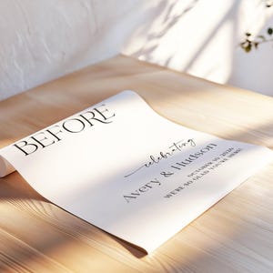 Pu&ograve; includere: Un foglio di carta bianco arrotolato con la parola "BEFORE" in un carattere nero grande ed elegante. La carta include anche il testo "celebrating Avery & Hudson October 10, 2026 WE'RE SO GLAD YOU'RE HERE!" su una superficie di legno.