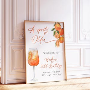 May include: A framed poster with a watercolor illustration of an Aperol Spritz cocktail and orange slices. The poster reads "A Spritz Older" and welcomes guests to Hailey's 30th birthday on August 25th, 20XX. The poster is 18x24 inches.