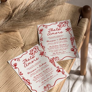 May include: Two white invitation cards with red text and illustrations. The text reads "Thato Amore" and "Join us for a rehearsal dinner in honor of Maisie & Lukas". The cards list the date, time, and location of the event. The illustrations include wine glasses, pizza slices, and peppers.
