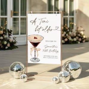 May include: A sign with the text "A Tini bit older" and "Welcome to Samantha's 30th Birthday". The sign features a cocktail illustration and is framed by a gold metal stand. Silver disco balls are in the foreground.