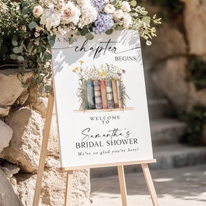 May include: A white sign on a wooden easel with the text "A New Chapter Begins" and "Welcome to Samantha's Bridal Shower". The sign is decorated with a floral arrangement and a watercolor illustration of books.