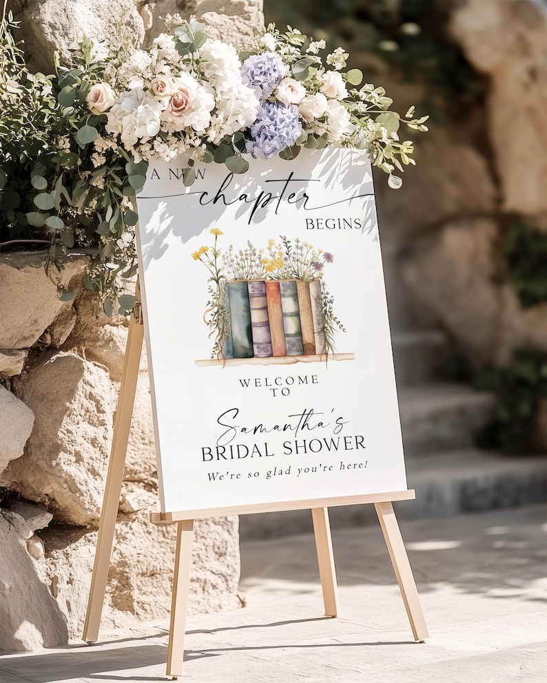 May include: A white sign on a wooden easel with the text "A New Chapter Begins" and "Welcome to Samantha's Bridal Shower". The sign is decorated with a floral arrangement and a watercolor illustration of books.