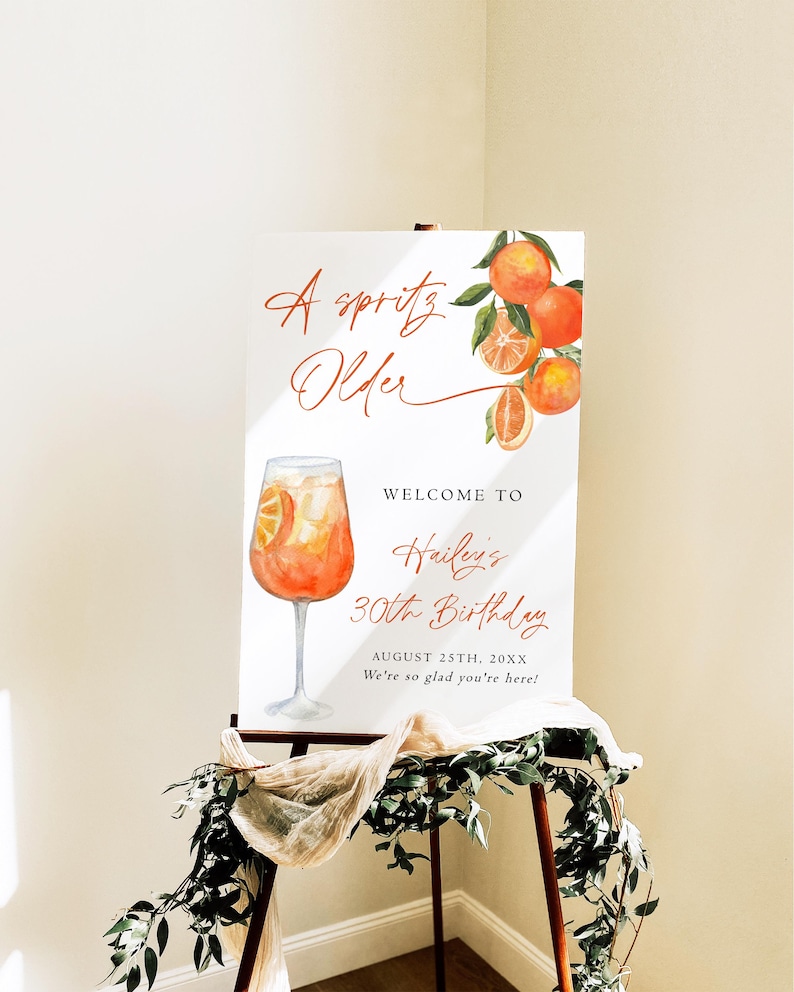 May include: A white sign on an easel with the text "A spritz Older" and "Welcome to Hailey's 30th Birthday". The sign features a watercolor illustration of an orange cocktail and oranges.