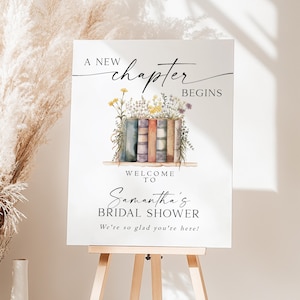 May include: A white sign on a wooden easel with the text "A New Chapter Begins" and "Welcome to Samantha's Bridal Shower". The sign features watercolor illustrations of books and flowers.