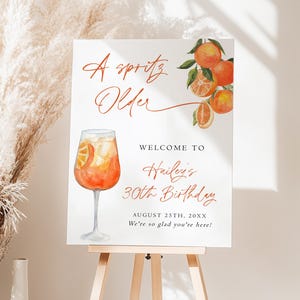 May include: A white sign on a wooden easel with the text "A spritz Older" in a cursive font. The sign also says "Welcome to Hailey's 30th Birthday" with the date and a welcoming message. An orange cocktail illustration is featured.