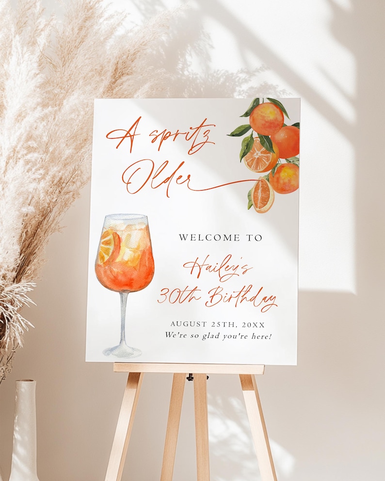 May include: A white sign on a wooden easel with the text "A spritz Older" in a cursive font. The sign also says "Welcome to Hailey's 30th Birthday" with the date and a welcoming message. An orange cocktail illustration is featured.