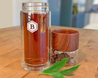 May include: A clear glass tea infuser bottle with a wooden lid and a white letter "B" on the side. The bottle is filled with brown tea and there are green tea leaves on a wooden surface.