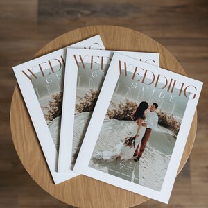 May include: Three white wedding guide magazines stacked on top of each other. The top magazine features a couple in a desert landscape with the text "Wedding Guide" in brown lettering.