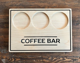 Puede incluir: Bandeja de barra de café de madera clara con tres recortes circulares. Las palabras "COFFEE BAR" están impresas en negro, centradas debajo de los recortes. La bandeja tiene un borde negro.