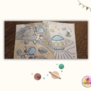 May include: A black and white coloring page featuring two astronauts in space suits, a flying saucer, and stars. The astronauts are wearing helmets and have big smiles. The flying saucer is shaped like a disc and has a window in the front. The stars are scattered across the page.