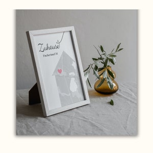 Puede incluir: Una impresi&oacute;n enmarcada en blanco con el texto "Zuhause" y "Fischerinsel 34" en escritura negra. La impresi&oacute;n presenta un mapa gris de un barrio con un coraz&oacute;n rojo marcando una ubicaci&oacute;n. La impresi&oacute;n se muestra en una superficie de lino blanco con un peque&ntilde;o jarr&oacute;n de hojas verdes.