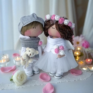 May include: Two handmade cloth dolls, a bride and groom, stand together. The groom doll wears a grey cap and scarf, holding a teddy bear. The bride doll wears a white dress and flower crown, holding a bouquet. Both dolls have rosy cheeks and angel wings.
